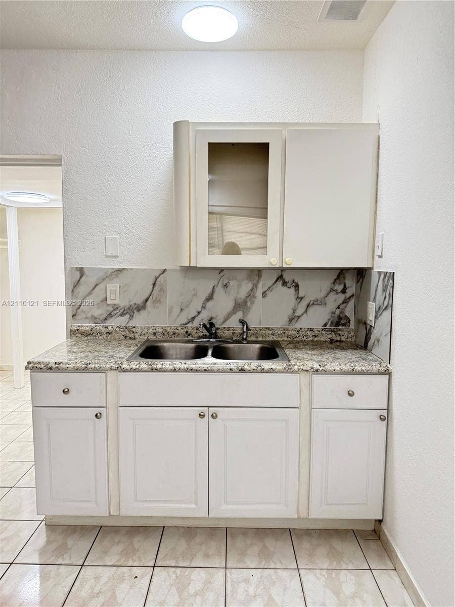 Kitchen area featuring white cabinetry, a dual-basin stainless steel sink, stone-finish countertop, decorative backsplash, and ceramic tile flooring