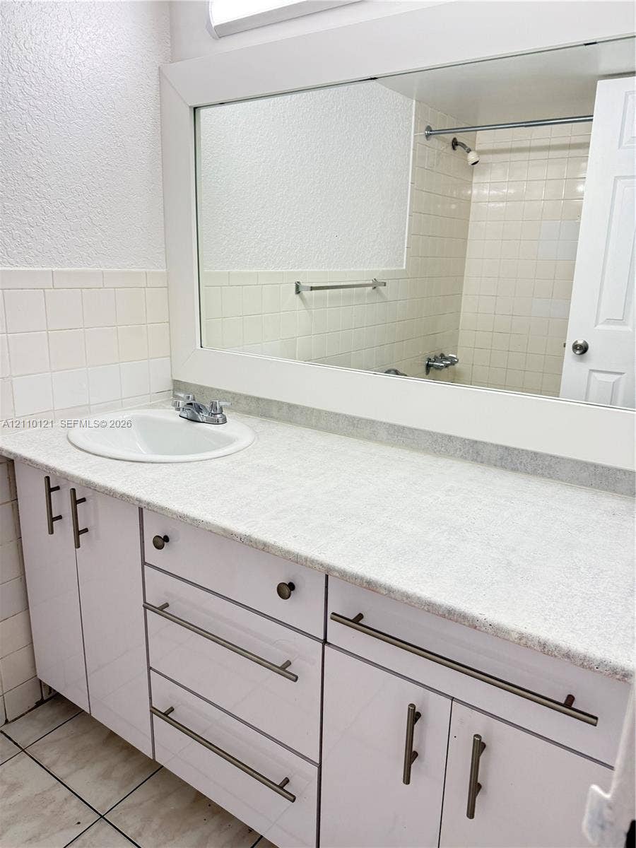 Bathroom vanity featuring white cabinetry, stone-finish countertop, integrated oval sink, modern bar-style hardware, and full-width wall mirror