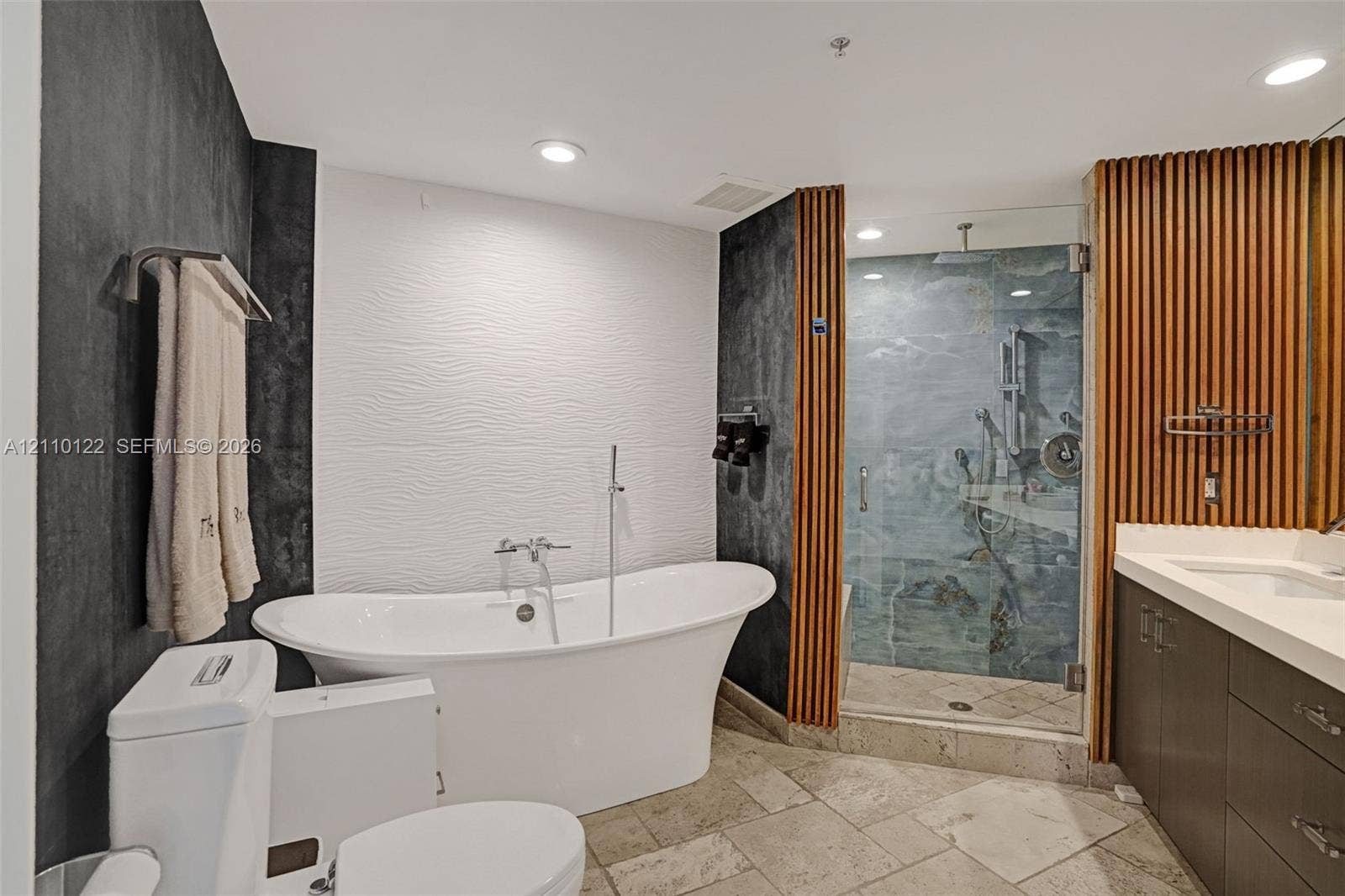 333 Las Olas Way, #2708, Fort Lauderdale, FL 33301 Freestanding soaking tub with a floor-mounted faucet, walk-in glass shower with stone tile interior, vertical wood-slat accent wall, stone-finish floor tiling, and dark-toned cabinetry with a solid-surface vanity countertop