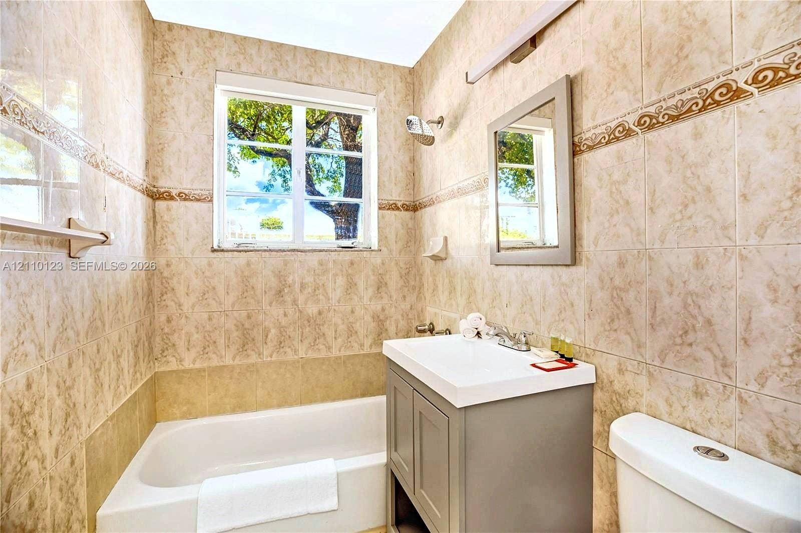 301 86th Street, #303-3, Miami Beach, Miami Beach, FL 33141 Full-height wall tiling with decorative border, bathtub-shower combination, grey vanity cabinet with integrated white sink, rectangular mirror, and bar lighting