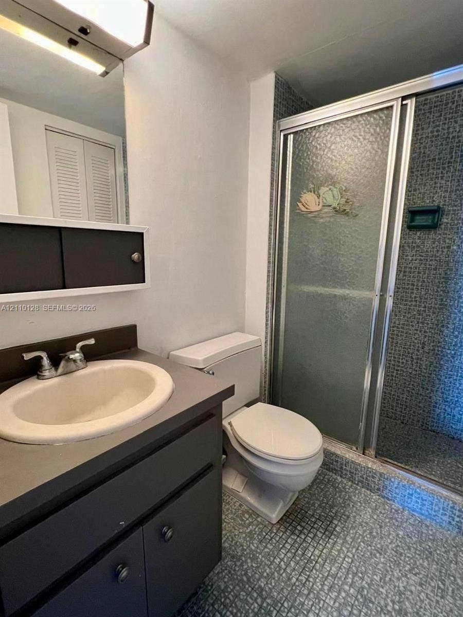 1351 NE Miami Gardens Drive, #1624E, Miami, FL 33179 Bathroom featuring a dark-finish vanity with a drop-in sink, wall-mounted medicine cabinet, modern linear vanity light, and a walk-in shower with textured glass doors and tiled surround