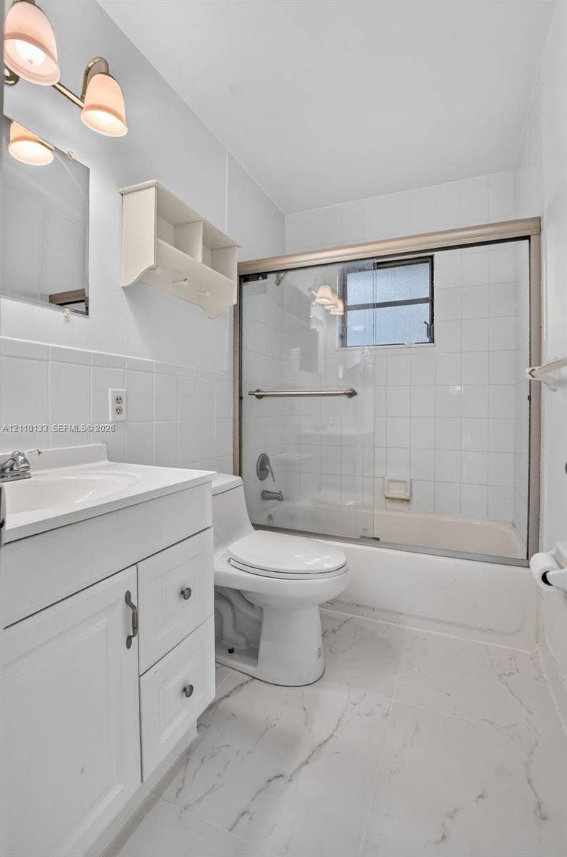 7890 Lago Del Mar Drive, #141, Boca Raton, Boca Raton, FL 33433 Bathroom featuring a white vanity with integrated sink, stone-patterned tile flooring, three-light wall-mounted fixture, and a glass-enclosed tub with shower surround