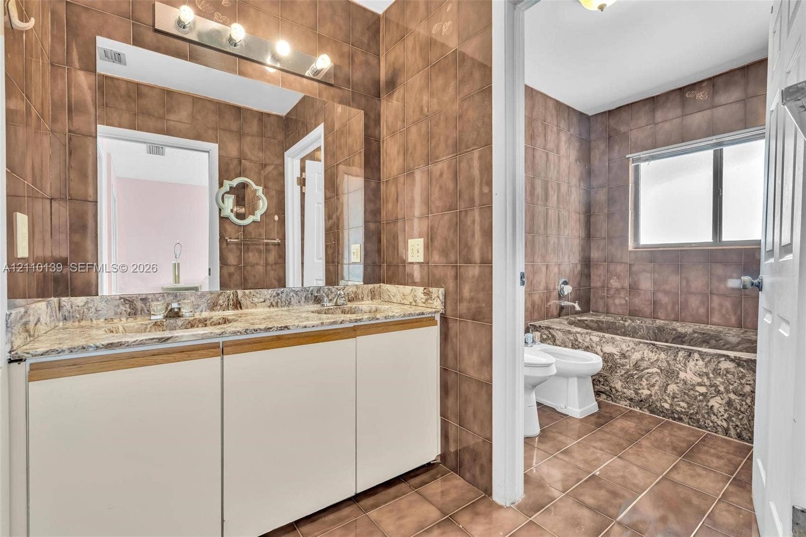 3601 NE 168th Street, #108, North Miami Beach, FL 33160 Bathroom featuring a dual-sink vanity with stone-finish countertop, full-wall tiling, a built-in stone-finish tub, and a bidet