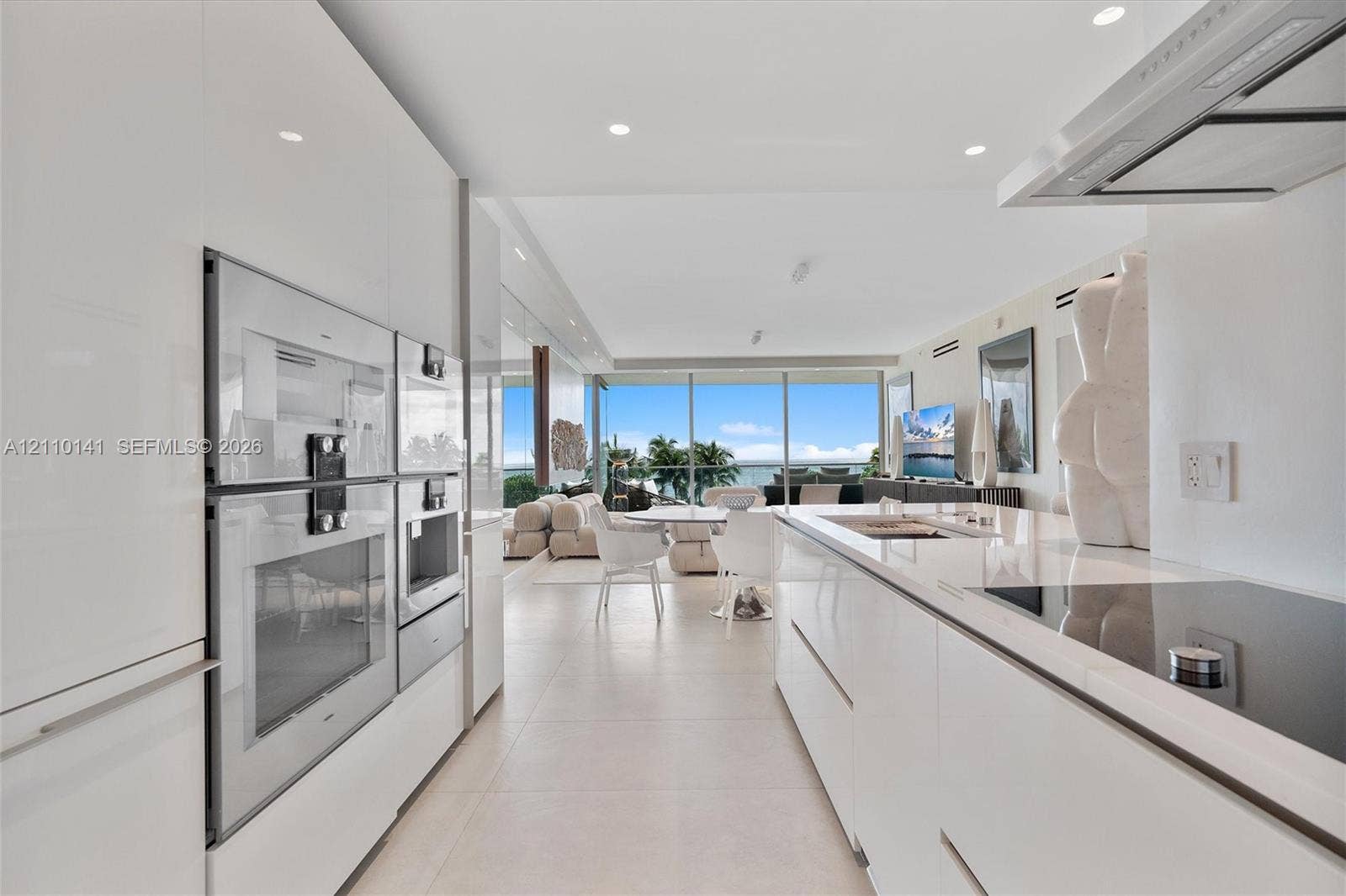 10203 Collins Avenue, #303, Bal Harbour, FL 33154 Contemporary kitchen featuring white high-gloss cabinetry, stainless steel built-in double wall ovens, a seamless solid-surface island countertop, integrated cooktop, and floor-to-ceiling glass paneling
