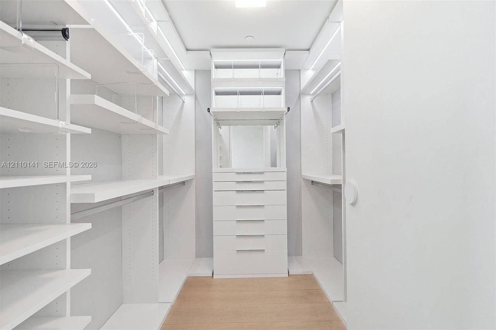 10203 Collins Avenue, #303, Bal Harbour, FL 33154 Custom walk-in closet featuring floor-to-ceiling white shelving, integrated LED accent lighting, a center stack of drawers, and wood-finish flooring
