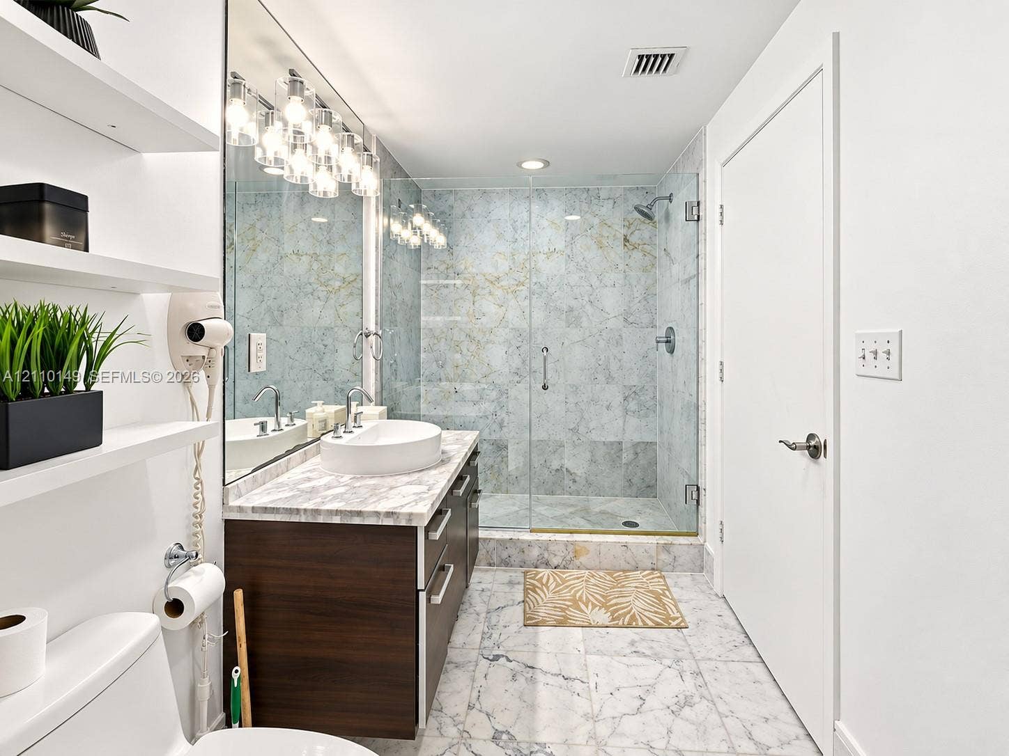Bathroom featuring a dark wood-finish vanity with a stone-top surface and vessel sink, walk-in shower with floor-to-ceiling tile surround and glass enclosure, wall-to-wall mirror, built-in shelving, and light-toned tile flooring