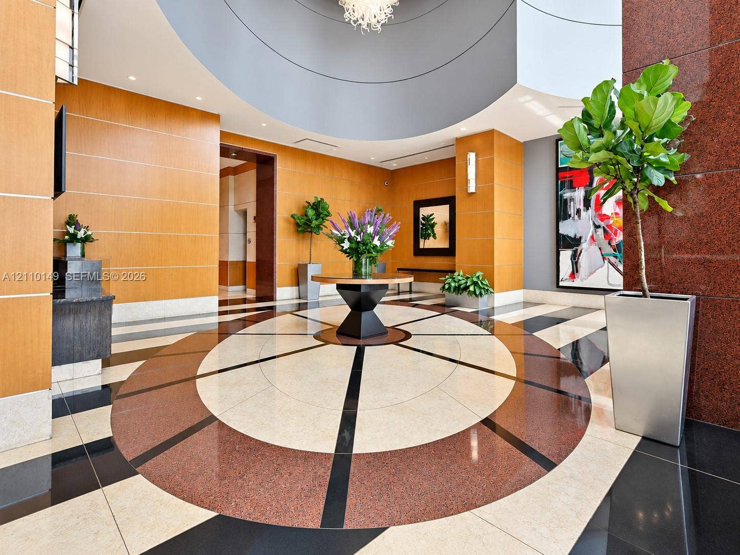 Grand foyer featuring geometric stone flooring, warm wood-paneled walls, a high circular ceiling, polished red stone accents, and a contemporary multi-arm chandelier