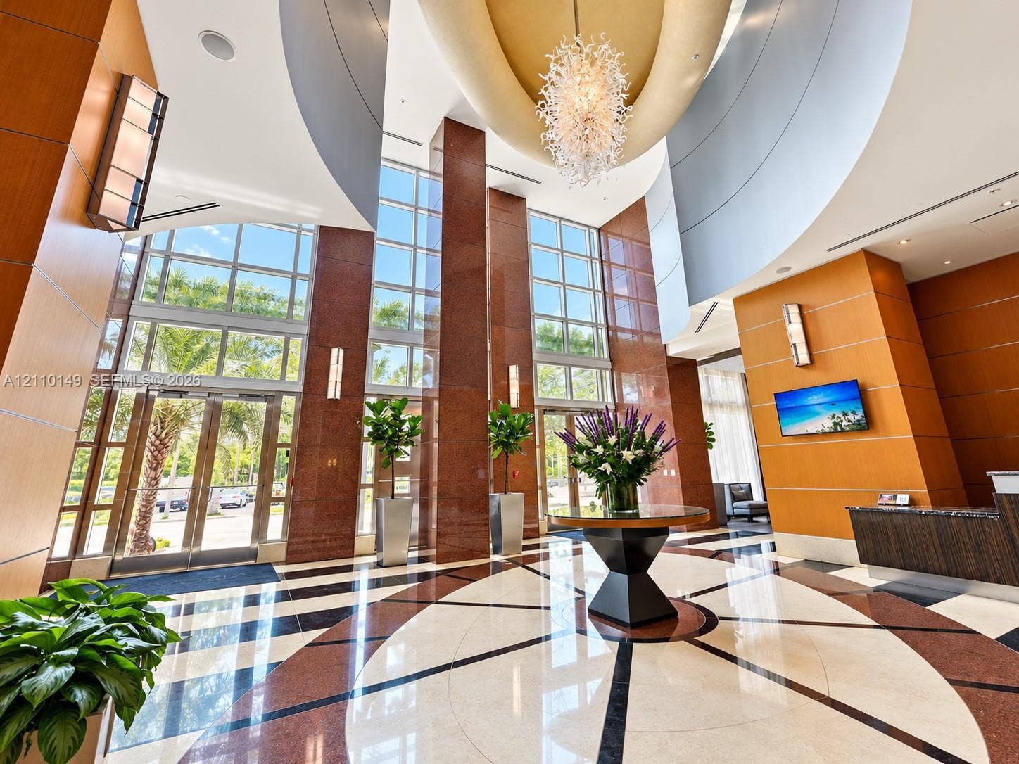 Grand atrium lobby featuring floor-to-ceiling windows, polished stone flooring with geometric inlays, wood-paneled wall accents, a multi-tiered circular tray ceiling, and a prominent chandelier