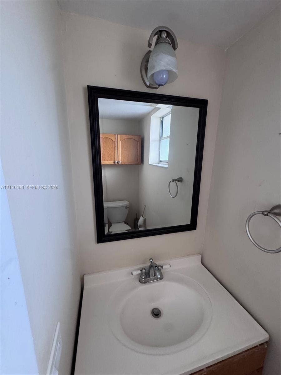 20111 NE 27th Court, #K212, Aventura, Aventura, FL 33180 Vanity featuring an integrated white sink, dual-handle metal faucet, wall-mounted light fixture, framed mirror, and brushed-metal towel ring