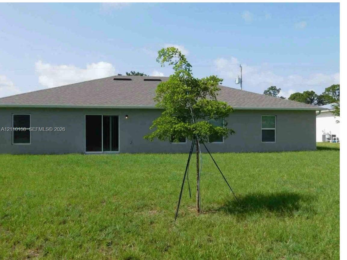 1743 SW Bellevue Avenue, Port St Lucie, FL 34953 Rear exterior elevation featuring stucco siding, a hip roof with asphalt shingles, sliding glass doors, and a grass-covered backyard