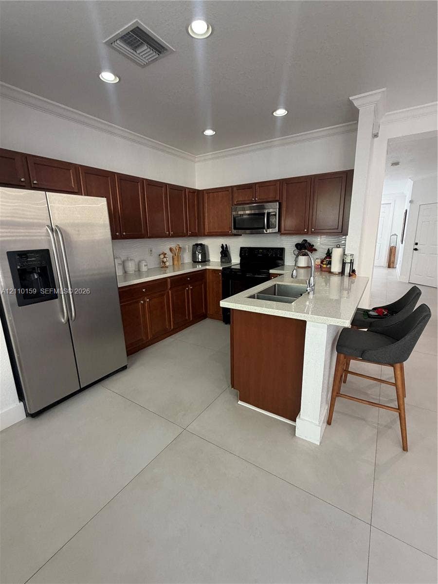 Kitchen featuring wood cabinetry, light-toned stone countertops, a center island with a dual-basin stainless steel sink, large-format tile flooring, and recessed ceiling lighting