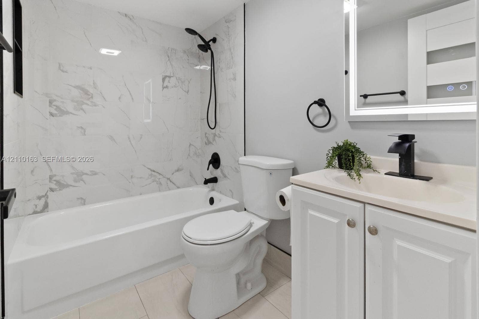 Bathroom featuring a soaking tub with full-height stone-look tile surround, vanity with an integrated sink, matte black faucet hardware, and a backlit mirror