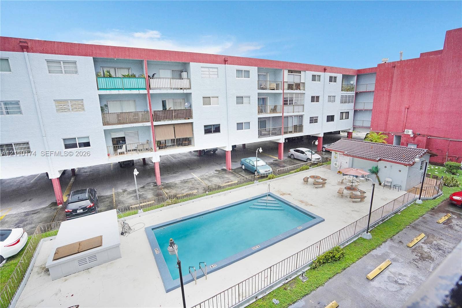 In-ground swimming pool with surrounding concrete patio, covered ground-level parking, and metal perimeter fencing