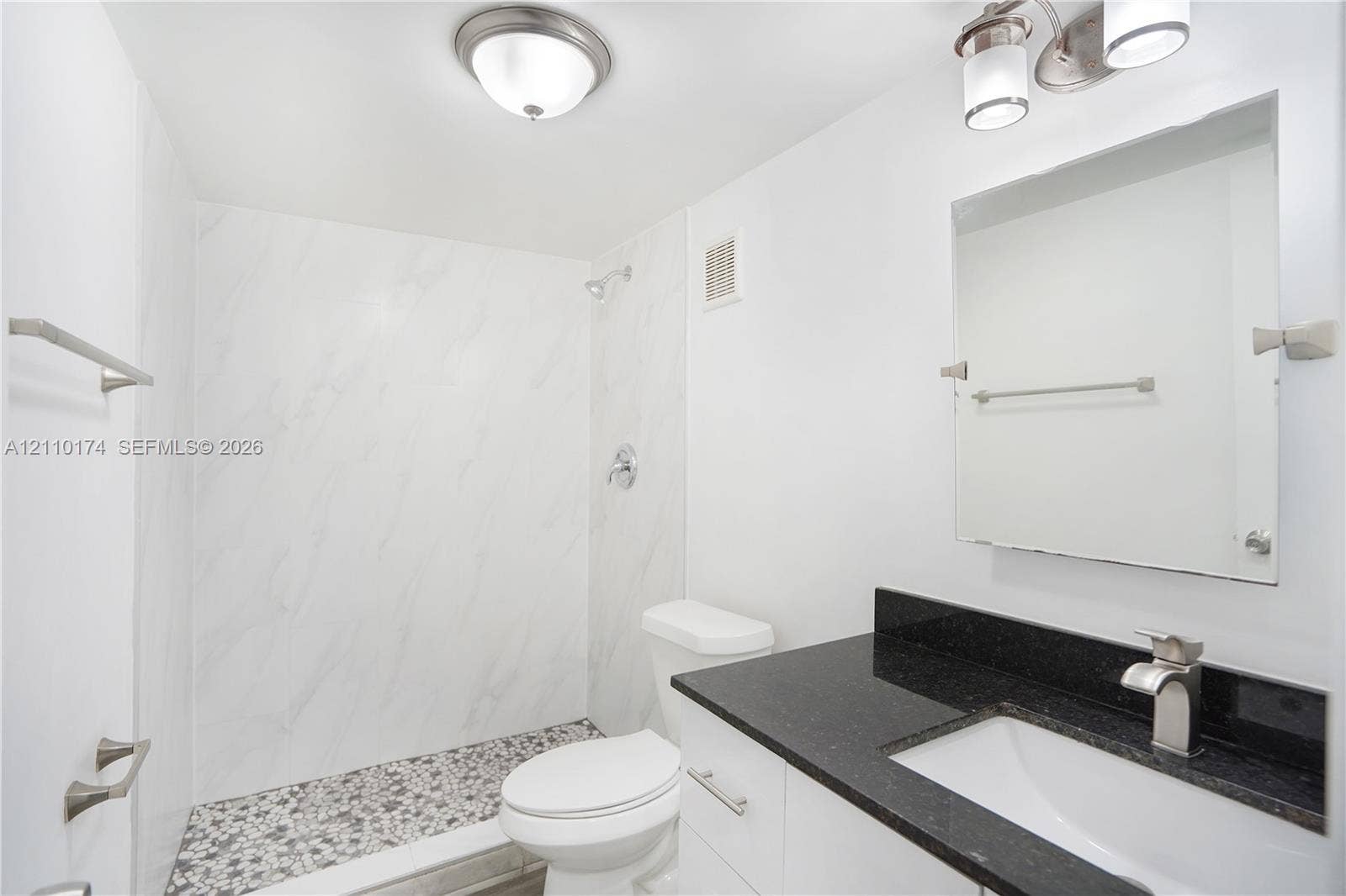 Full-height shower featuring large-format stone-pattern wall tiles and a decorative pebble-style basin floor, vanity with a solid-surface countertop and undermount sink, and wall-mounted vanity mirror with integrated lighting