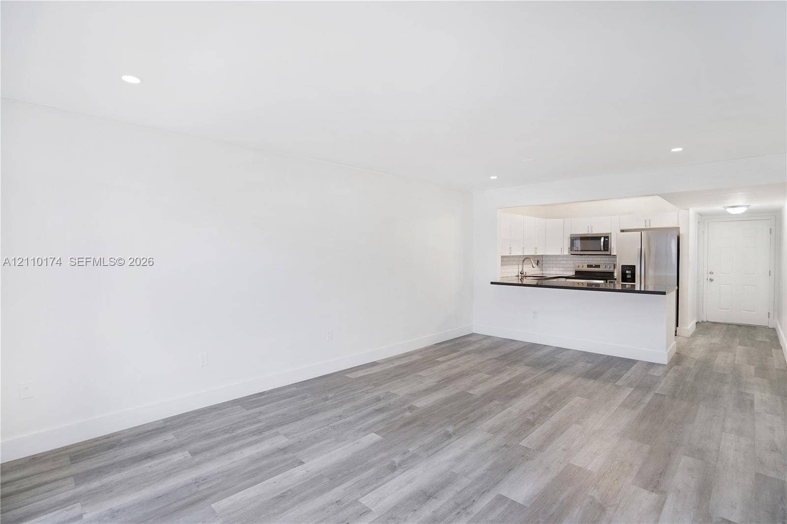 Open-concept interior featuring wood-finish flooring, recessed lighting, and a kitchen with white cabinetry, stainless steel appliances, and a dark-toned countertop breakfast bar