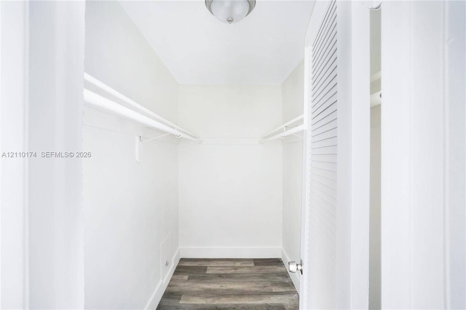 Storage area featuring wood-finish flooring, dual-level wall-mounted shelving, and a louvered door