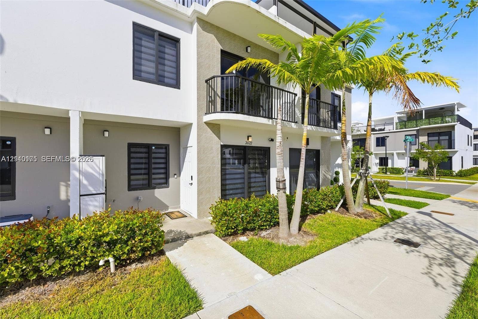 26122 SW 145th Court, #26122, Homestead, Homestead, FL 33032 Two-story exterior featuring a stucco facade, second-floor balcony with black metal railing, concrete entry path, and mature palm landscaping