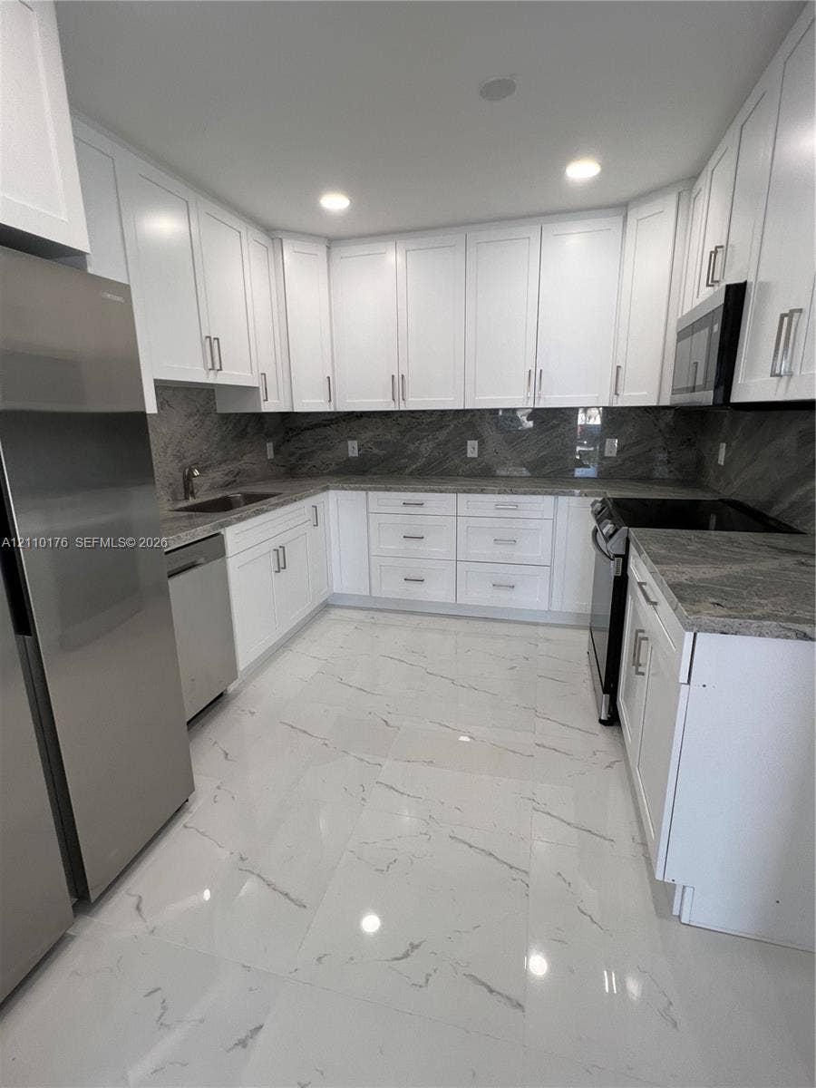 Kitchen featuring white shaker-style cabinetry, stone-finish countertops, full-height stone-finish backsplash, stainless steel appliances, and large-format porcelain tile flooring