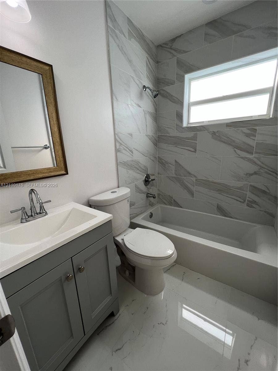 Bathroom featuring gray shaker-style vanity cabinetry, white integrated sink countertop, large-format tile flooring, and tub-shower combination with full-height wall tiling