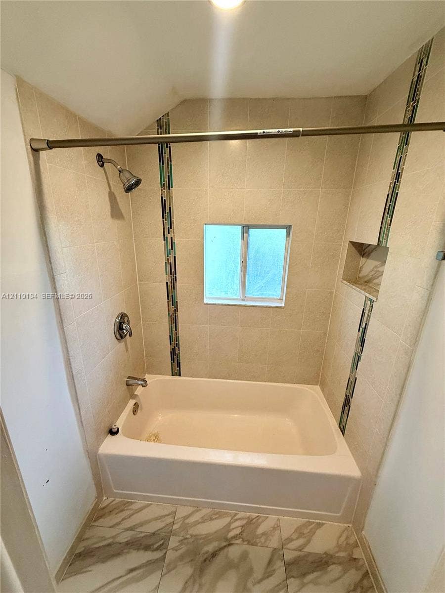 Bathtub and shower combination featuring neutral tile surround with decorative glass mosaic vertical accents, built-in recessed storage niche, and stone-finish floor tile