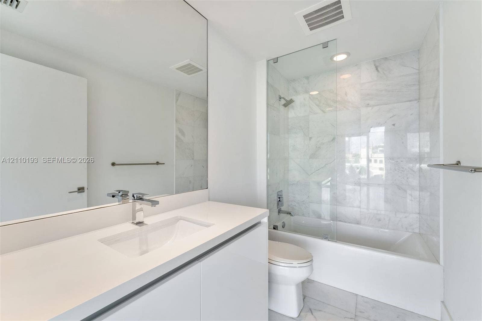 Modern vanity with a rectangular basin, expansive wall-to-wall mirror, shower-tub combination, stone-tile surround, and frameless glass panel
