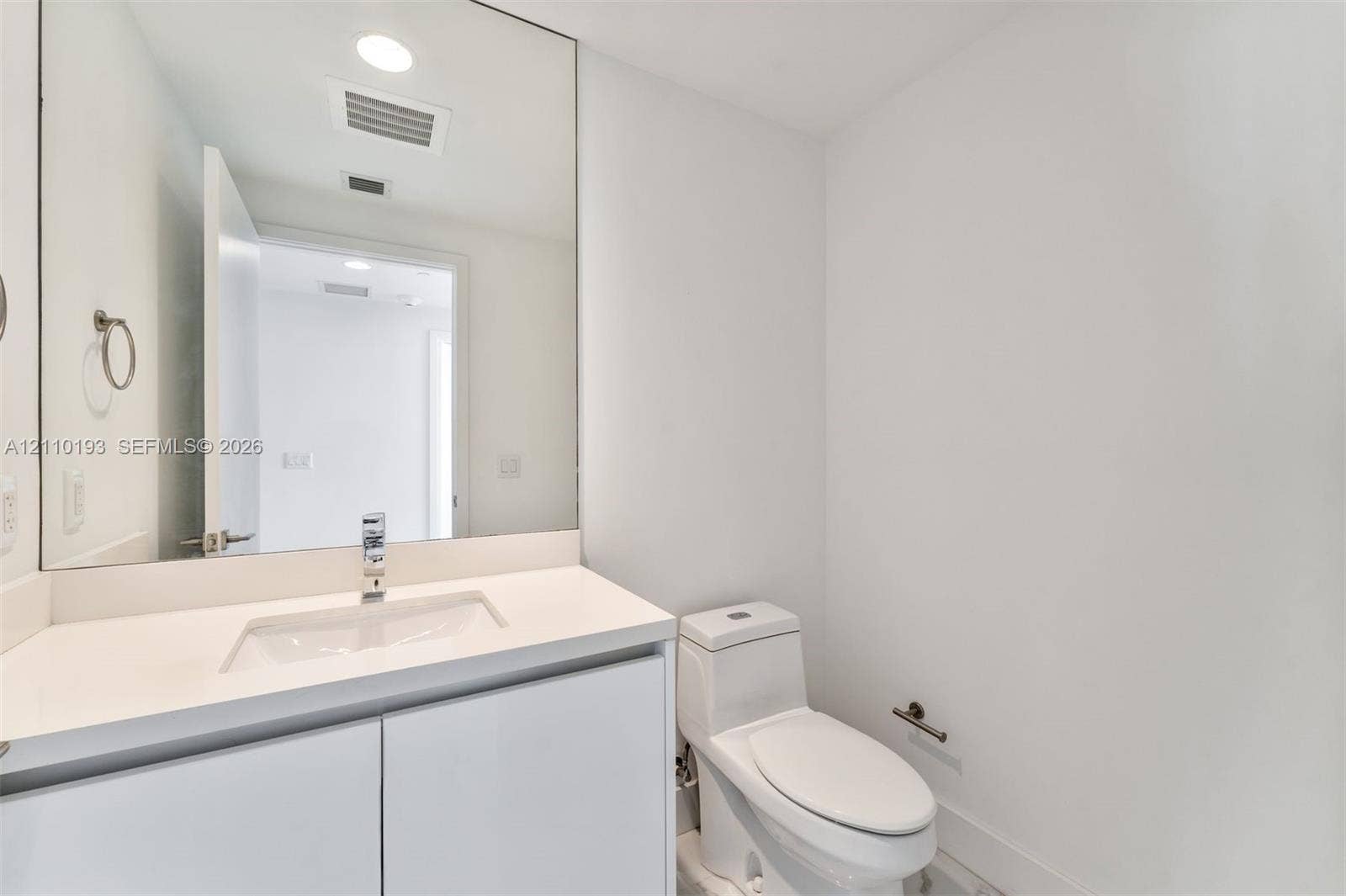 White vanity with solid-surface countertop, rectangular integrated sink, chrome single-handle faucet, full-width mirror, and dual-flush toilet
