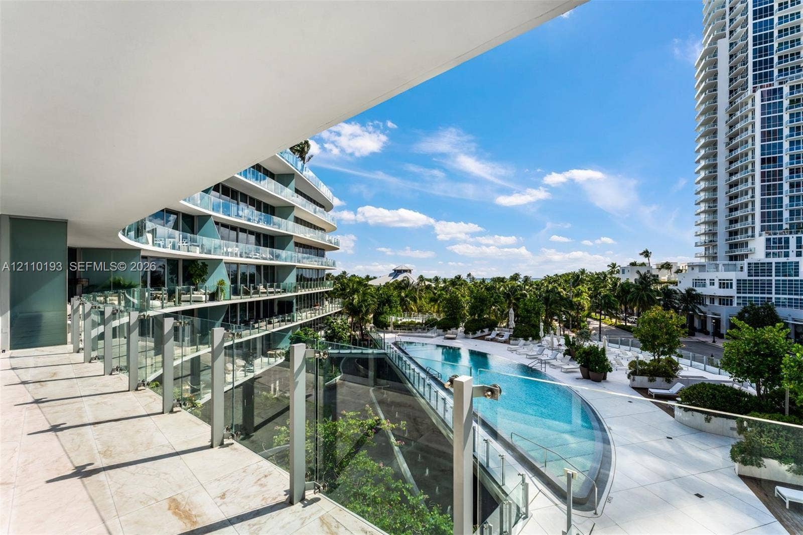 Covered terrace featuring stone tile flooring and glass panel railings, overlooking a swimming pool with a stone deck, mature palm trees, and structural landscape elements
