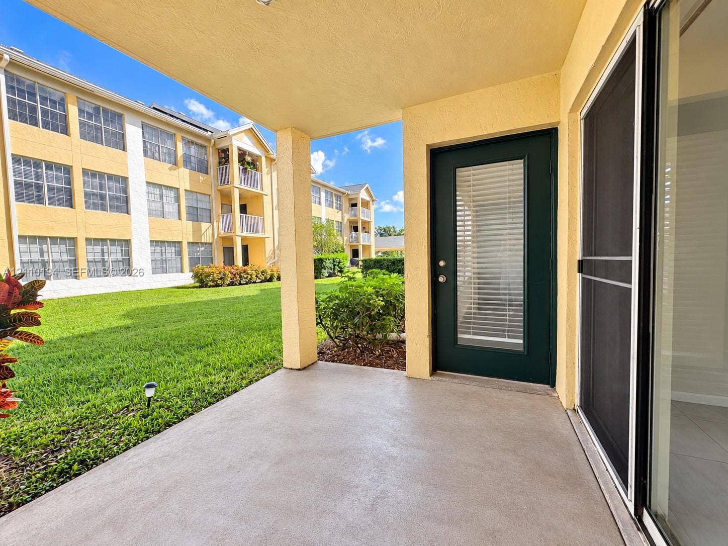 6331 La Costa Drive, #A, Boca Raton, Boca Raton, FL 33433 Covered concrete patio featuring a solid-color door with inset window, sliding glass door with mesh screen, and exterior lighting