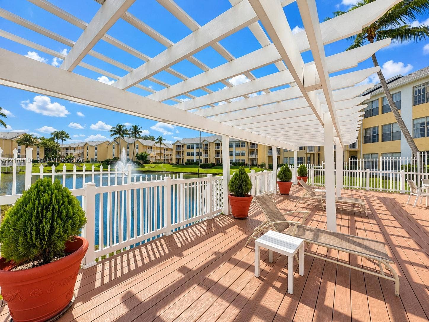 6331 La Costa Drive, #A, Boca Raton, Boca Raton, FL 33433 Expansive wood-tone deck featuring a white overhead pergola and white picket-style perimeter fencing overlooking a pond with a built-in fountain