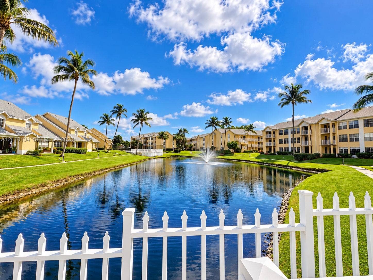 6331 La Costa Drive, #A, Boca Raton, Boca Raton, FL 33433 Community pond featuring a central fountain, manicured lawn, white picket fencing, and surrounding palm trees