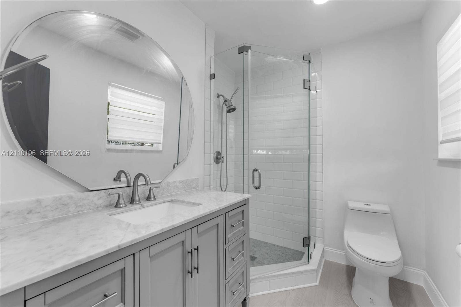 4400 SW 89th Avenue, #4400, Miami, Miami, FL 33165 Bathroom featuring a gray vanity with a neutral stone countertop, an oversized circular mirror, a glass-enclosed shower with subway tile surround, and wood-finish flooring