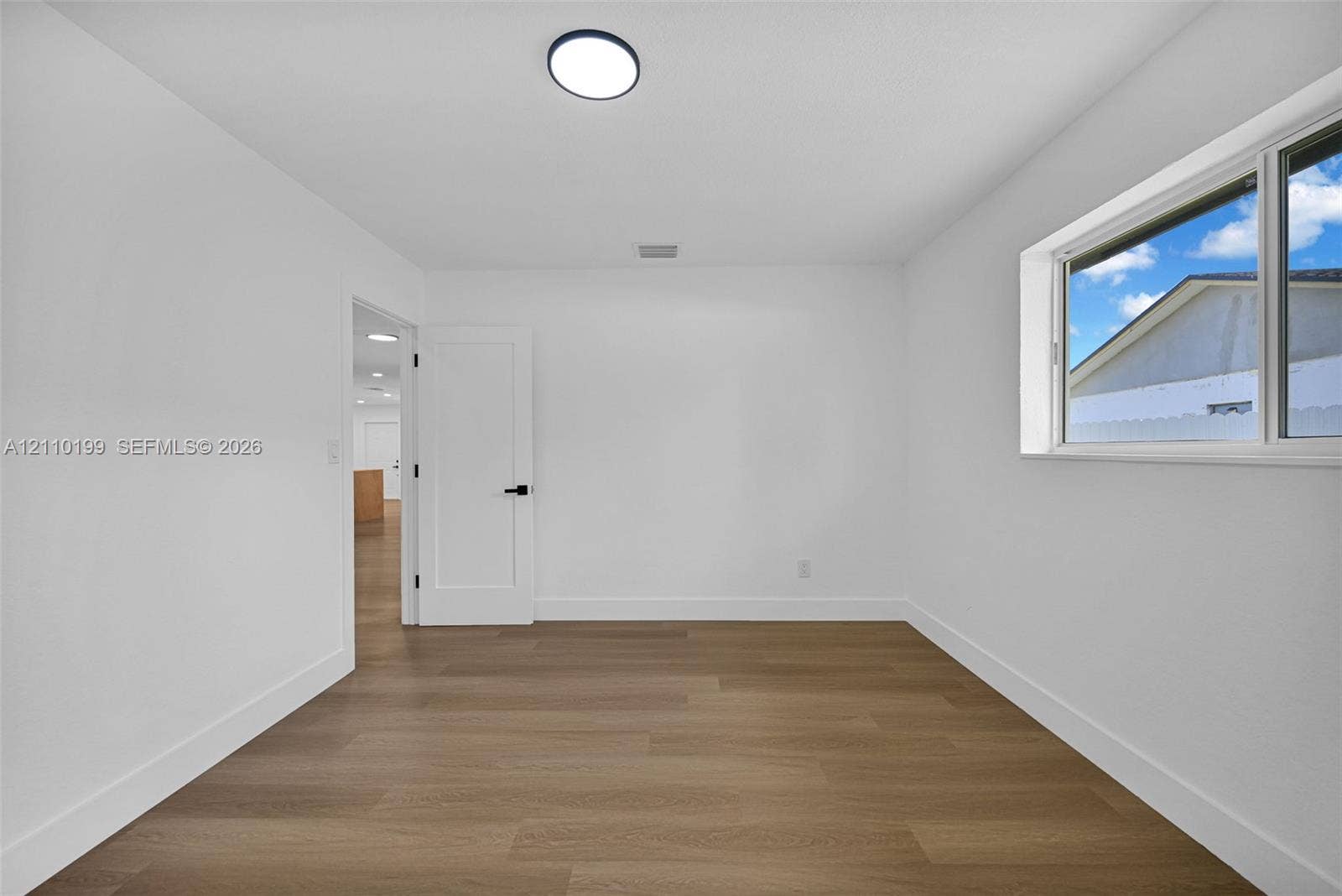 10741 SW 165th Street, Miami, FL 33157 Room featuring wood-finish flooring, crisp white walls, a dual-pane sliding window, a modern flat-panel door with matte black hardware, and a flush-mount LED light fixture