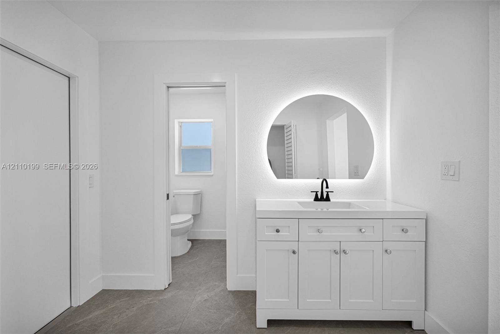 10741 SW 165th Street, Miami, FL 33157 White vanity featuring a solid-surface countertop with an integrated rectangular sink and matte black faucet, illuminated semi-circular wall mirror, and neutral stone-finish tile flooring