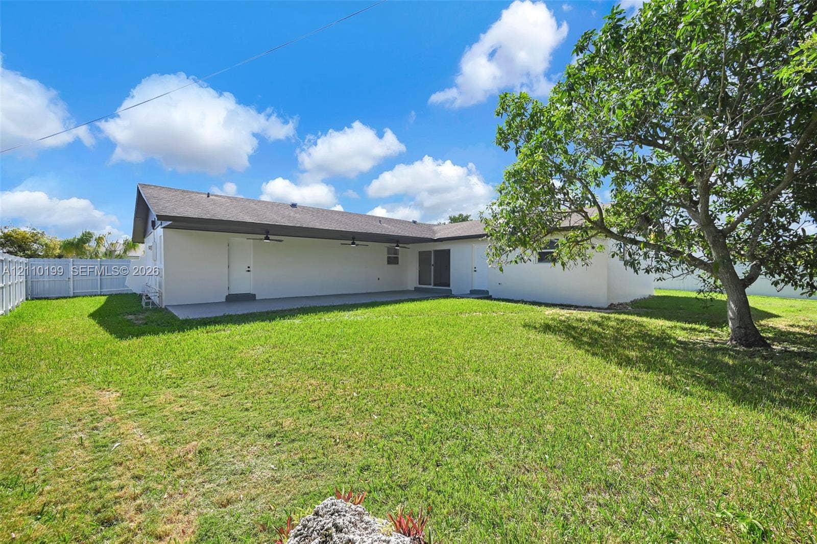 10741 SW 165th Street, Miami, FL 33157 Expansive backyard featuring a manicured lawn, mature trees, a covered patio with ceiling fans, and a white privacy fence