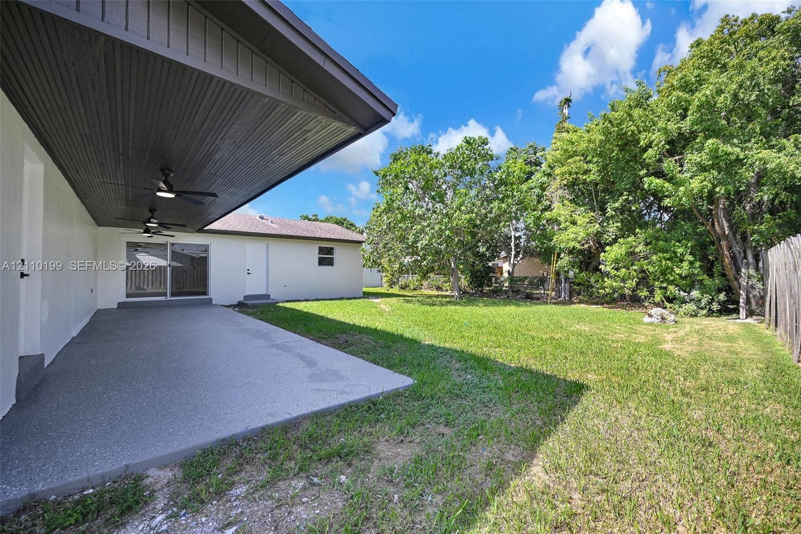 10741 SW 165th Street, Miami, FL 33157 Covered concrete patio with beadboard ceiling and dual built-in ceiling fans, featuring a landscaped backyard with mature trees and perimeter wood fencing