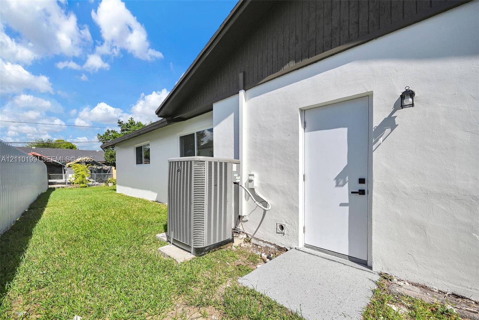 10741 SW 165th Street, Miami, FL 33157 Exterior side yard featuring white stucco siding, dark wood-finish eaves, an exterior access door with matte black hardware, a wall-mounted sconce, and a central air conditioning unit