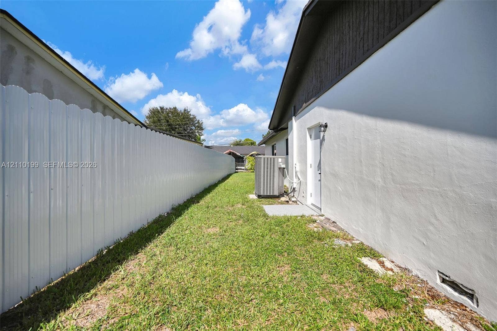 10741 SW 165th Street, Miami, FL 33157 Side yard featuring a white corrugated metal fence, grass lawn, and exterior wall-mounted lighting