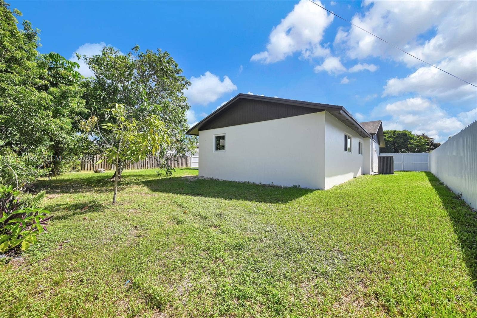 10741 SW 165th Street, Miami, FL 33157 Exterior side profile featuring light-toned stucco siding, a low-pitched roof with dark-toned gable end, and a spacious grass yard enclosed by a corrugated metal fence
