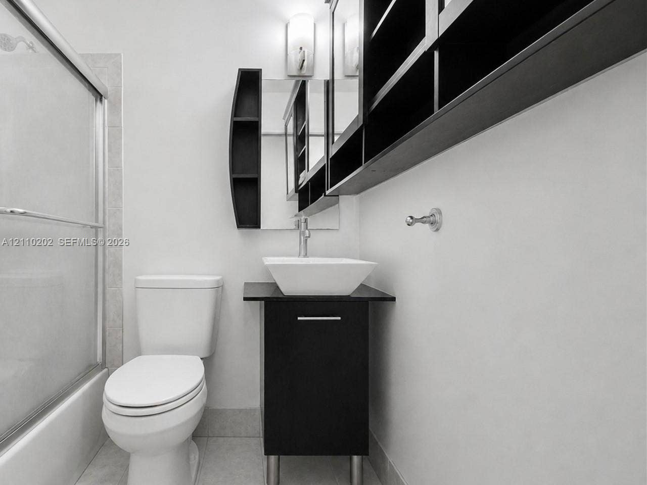 Modern vanity featuring a dark-finish base, square vessel sink, and chrome faucet, accompanied by built-in wall-mounted cabinetry and a glass-enclosed tub shower