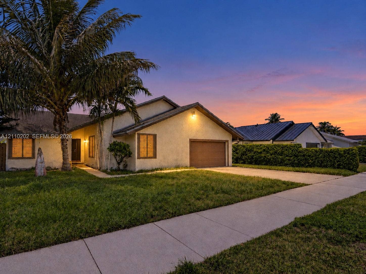 Single-story exterior featuring light-toned stucco siding, an attached garage, and mature palm trees