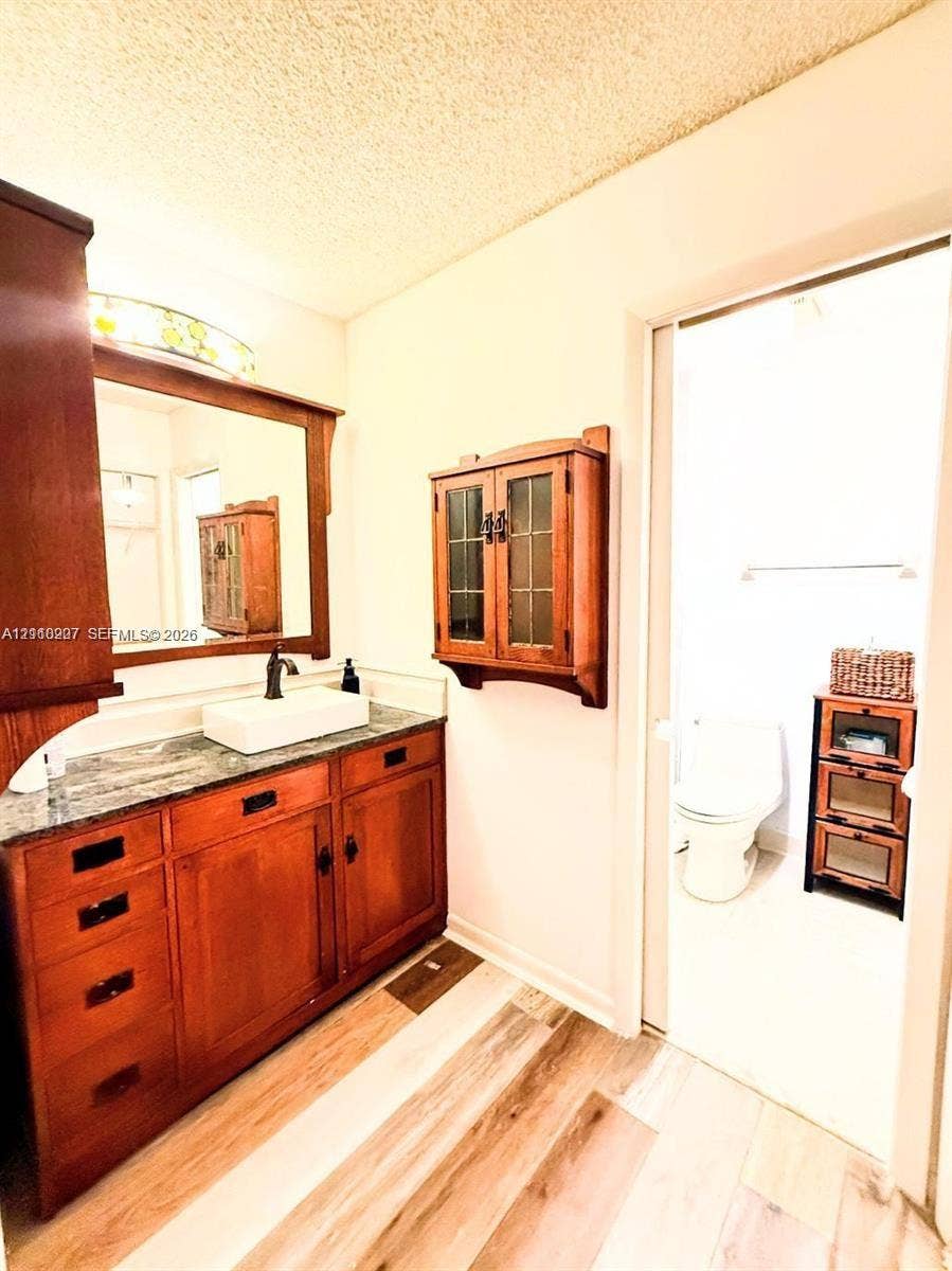 766 NW 24th Avenue, Delray Beach, FL 33445 Bathroom featuring stained wood cabinetry, rectangular vessel sink, stone-finish countertop, wall-mounted wood-framed storage, and wood-finish flooring