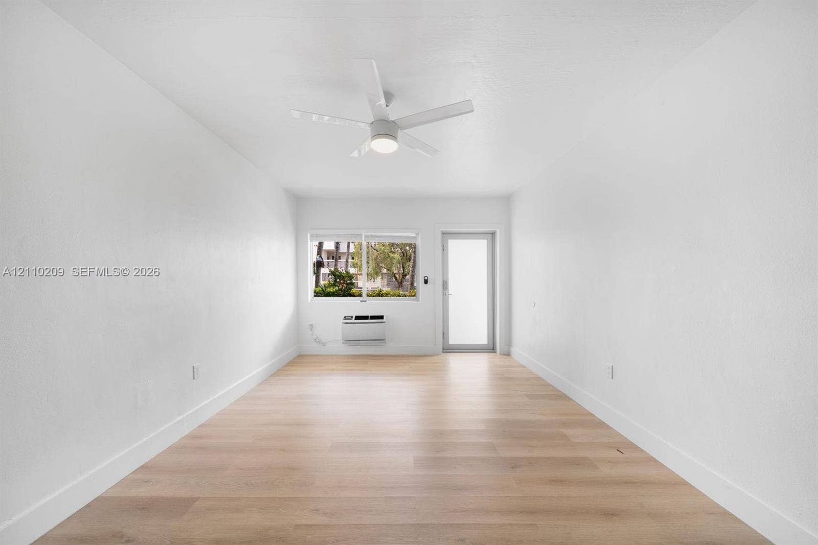 Bright interior featuring wood-finish flooring, a contemporary ceiling fan, a large window, and an integrated wall-mounted climate control unit