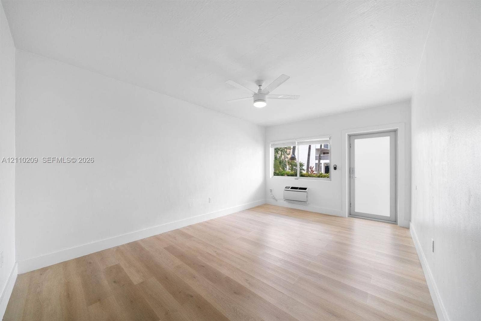 Room featuring wood-finish flooring, a white ceiling fan with integrated lighting, a wall-mounted air conditioning unit, and a glass-paneled exterior door