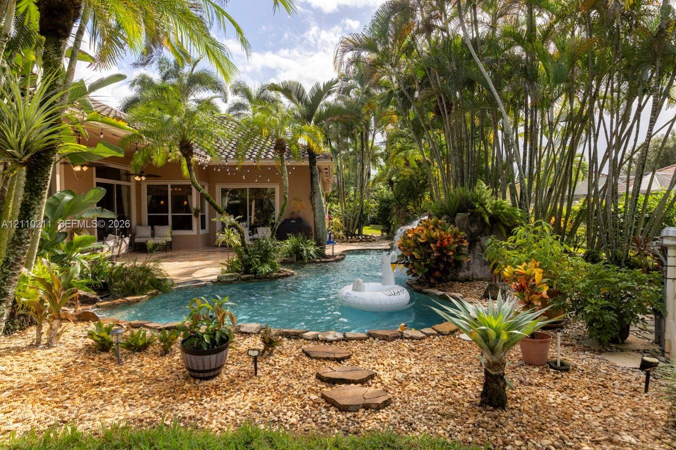 Freeform swimming pool featuring a stone-accented water feature, concrete patio, pebble-style landscaping, and mature palm trees