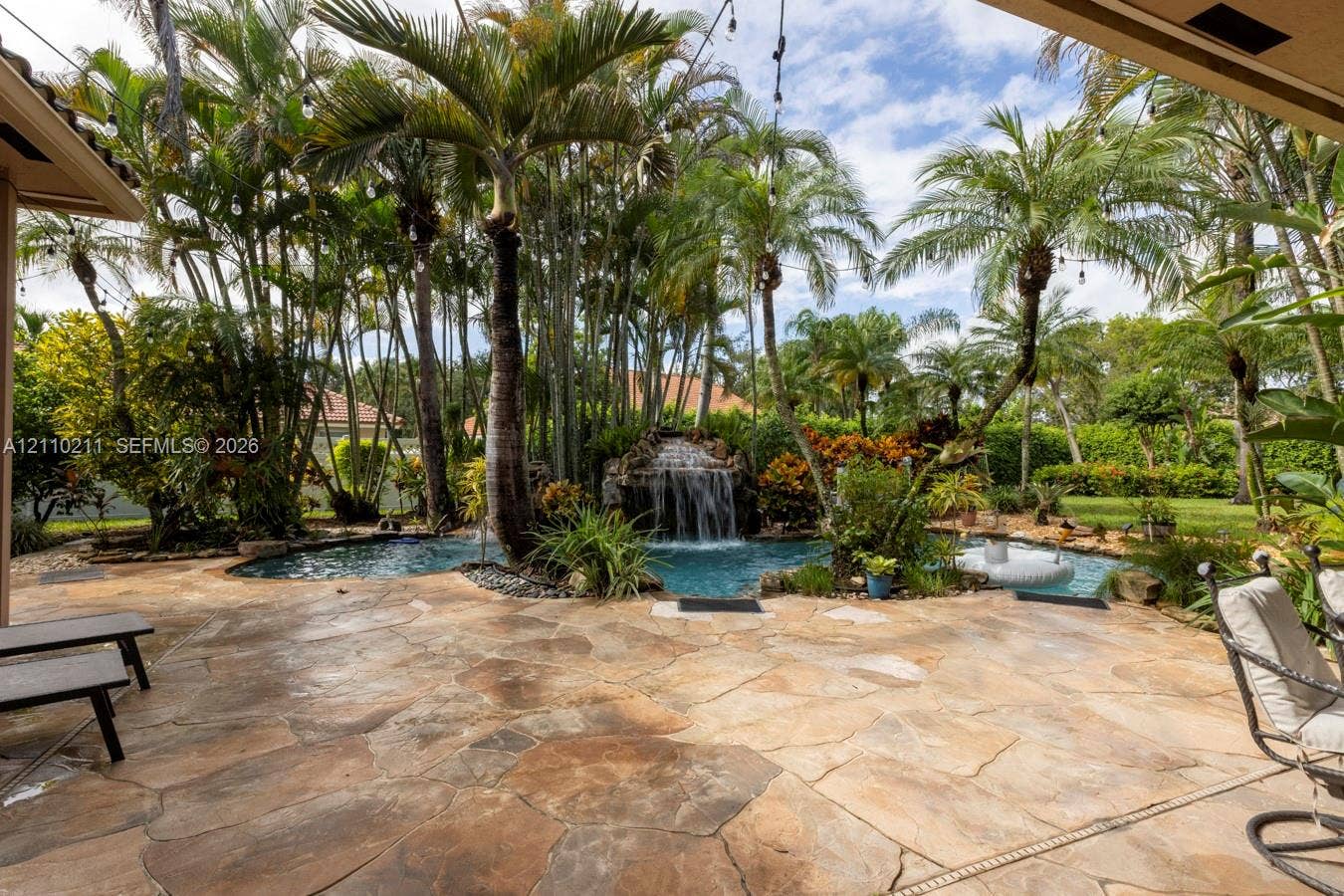 Flagstone patio area featuring a free-form pool with a rock waterfall feature, mature palm trees, and lush tropical landscaping