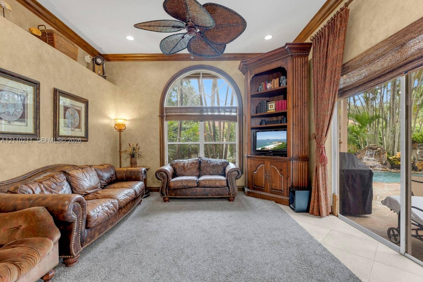 Living area featuring crown molding, arched window, built-in wooden corner cabinetry, palm-style ceiling fan, and sliding glass door