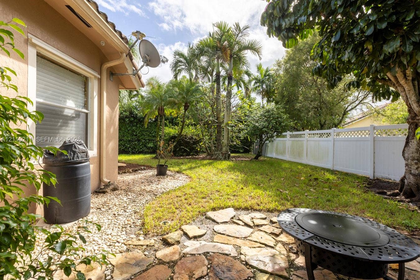 Private backyard featuring a stone patio, manicured lawn, perimeter white vinyl fencing, and mature palm trees