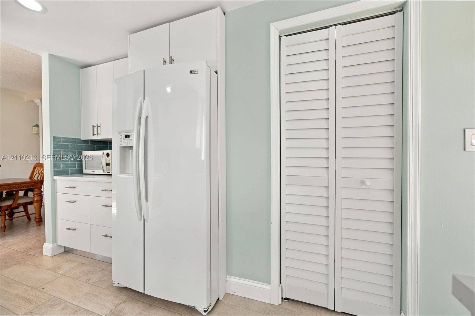 White shaker-style cabinetry with overhead storage, white side-by-side refrigerator, white louvered-style closet doors, blue tile backsplash, and tile flooring