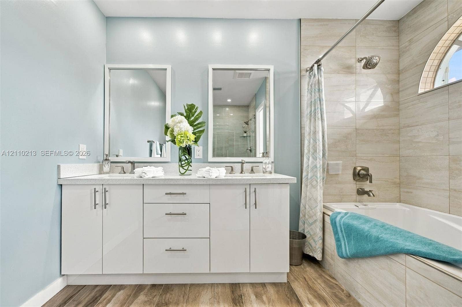 Dual-sink vanity with white cabinetry and light-toned solid surface countertop, wood-finish flooring, full-height tile shower surround, and semi-circular window
