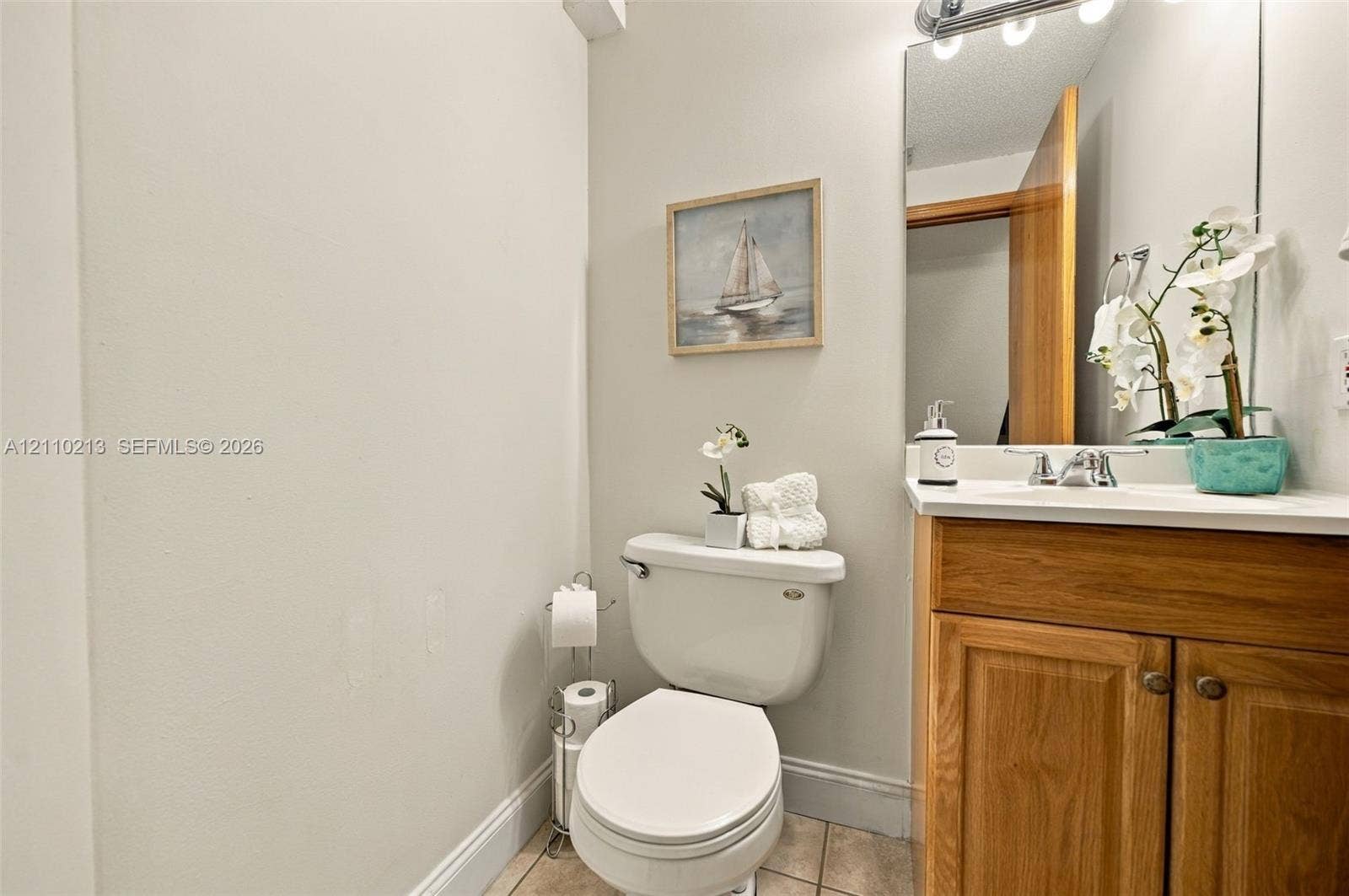 Wood-finish cabinetry, solid-surface countertop with integrated basin, polished chrome faucet, full-width vanity mirror, and ceramic tile flooring